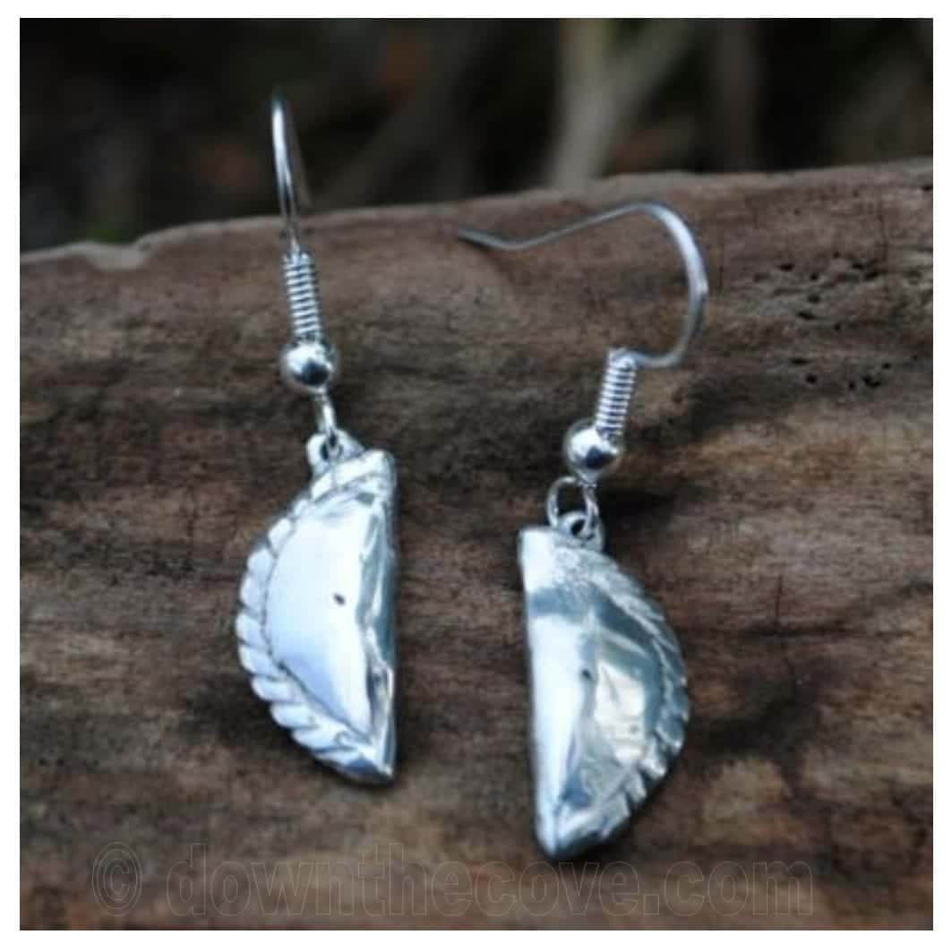Pair of Cornish pasty pewter earrings with crimped edges, hanging on silver hooks against driftwood