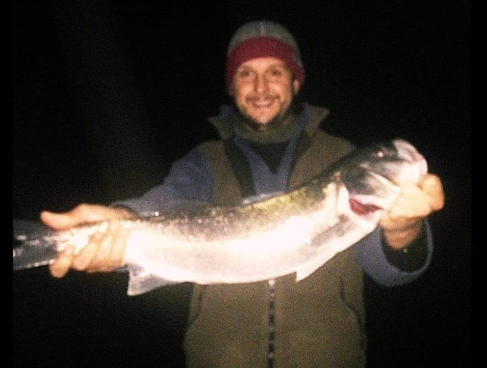 Smiling angler in a woolly hat holding a large sea bass at night